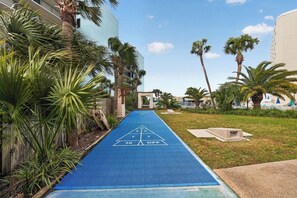 Una cancha de shuffleboard azul con un objetivo triangular, rodeada de palmeras y un edificio.