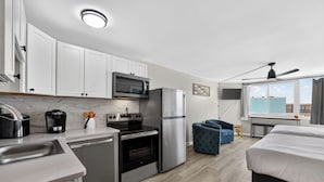 A modern kitchen with white cabinets, a microwave, a refrigerator, an oven, and a coffee maker. There is a bed with white bedding and a blue chair. - Beachfront Studio - Ocean View • Pool • Sleeps 4 (Panama City Beach)