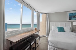 Un dormitorio con un ventanal grande, una cama, un escritorio y dos sillas, y vista al océano.