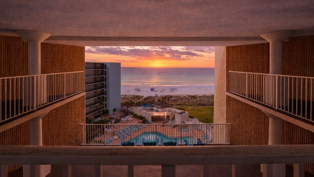 A balcony with a view of a sunset over the ocean, a pool, and a building with vertical wooden panels. - Beachfront Studio - Ocean View • Pool • Sleeps 4 (Panama City Beach)