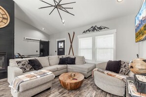 A modern living room with a sectional sofa, a round wooden coffee table, and a unique ceiling light fixture. - 👉 Level Up Lodge | Arcade + Hot Tub + Playground (Island Park)
