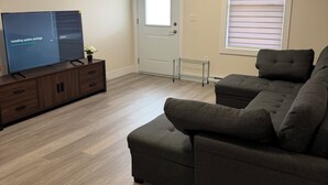 Een woonkamer met een flatscreen televisie op een houten kast, een grijze bank en een deur met een raam.