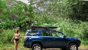 Un SUV azul estacionado sobre un camino de tierra con abundante vegetación de fondo.