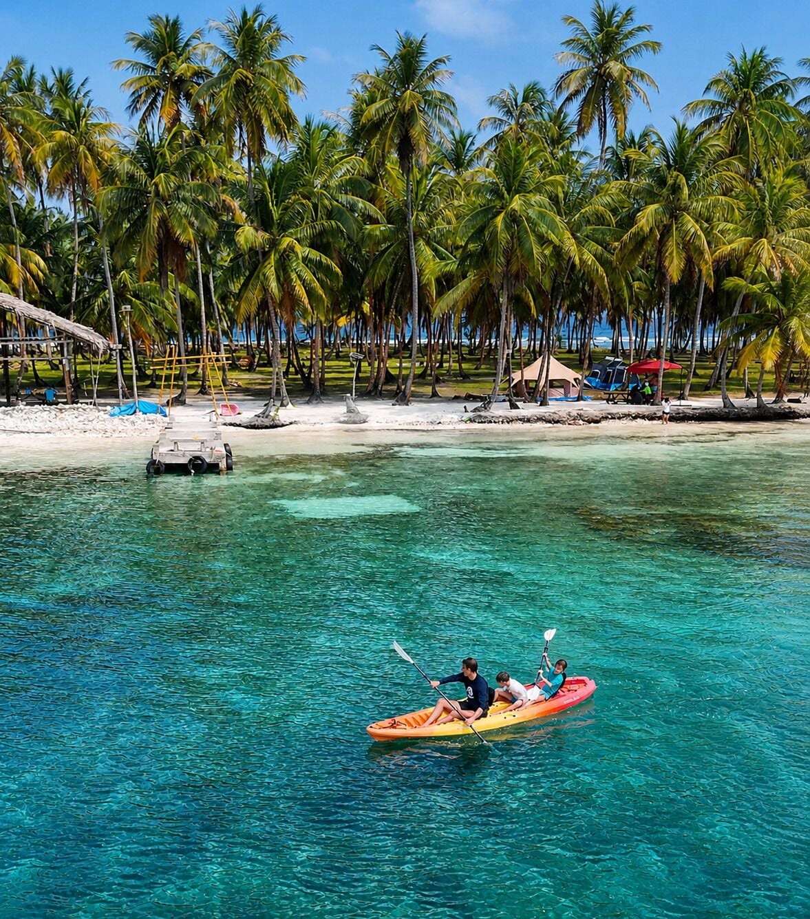 A clear tropical beach with a kayak in the water, palm trees, and a sandy shore with beach chairs.