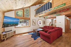A spacious living room with a red sofa, wooden flooring, a large window with a scenic view, and a loft area with wooden beams and signs. - Ruby Retreat by Tahoe Getaways - Pet Friendly 3 BR in Tahoe Donner (Truckee)