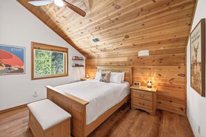 Quarto com painéis de madeira, uma cama, mesas de cabeceira, um banquinho e um ventilador de teto.