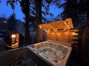 A hot tub with a wooden deck and string lights, surrounded by trees.