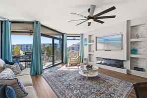 A modern living room with a large TV, a ceiling fan, a sofa with patterned cushions, a coffee table, and a view of the city through large windows. - Intracoastal Waterfront Escape w/ Dock, Hot Tub & Walk to Beach (New Smyrna Beach)