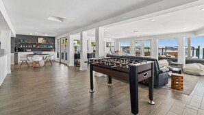 A spacious living area with a foosball table, a kitchen with a bar, and large windows offering a view of the water.