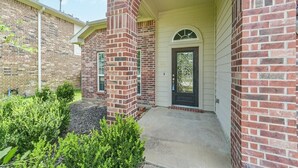 A brick building with a covered entrance, a door with a glass panel, and a small garden area. - Katy Brick Haven (Katy)