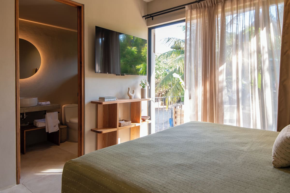 Un dormitorio con cama, televisor montado en la pared, baño con inodoro y lavabo, y un balcón con cortinas.