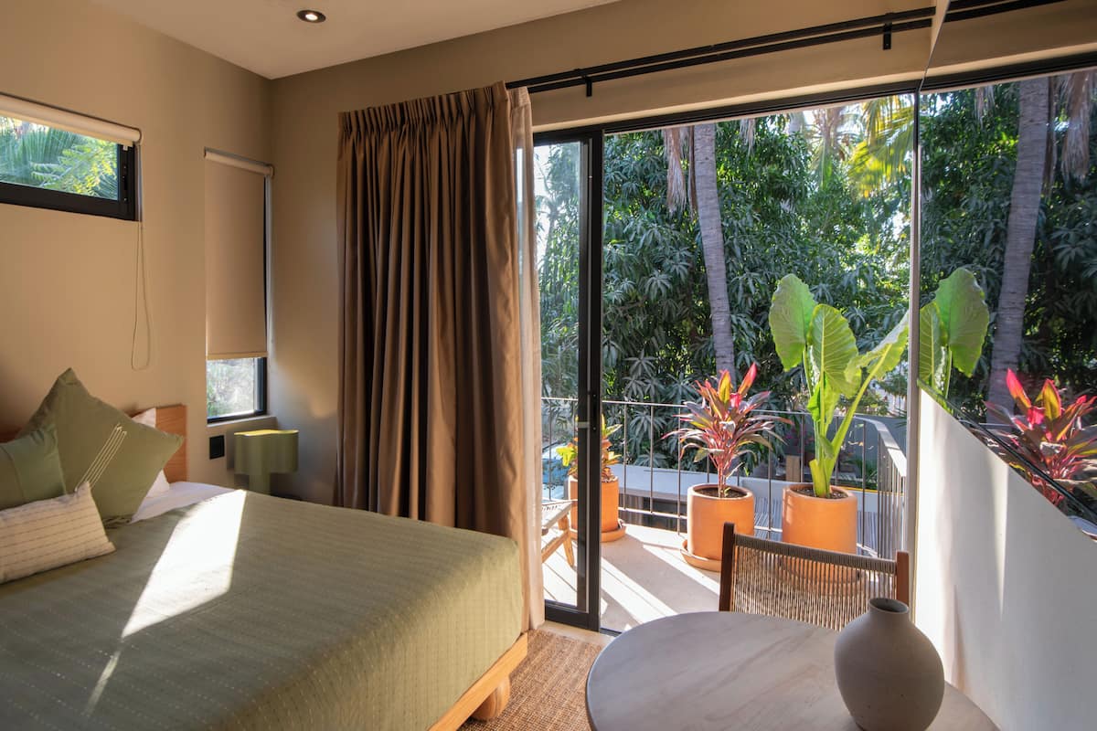 Un dormitorio con cama, ventana con cortinas, mesita de noche y vistas a un balcón con plantas en macetas.
