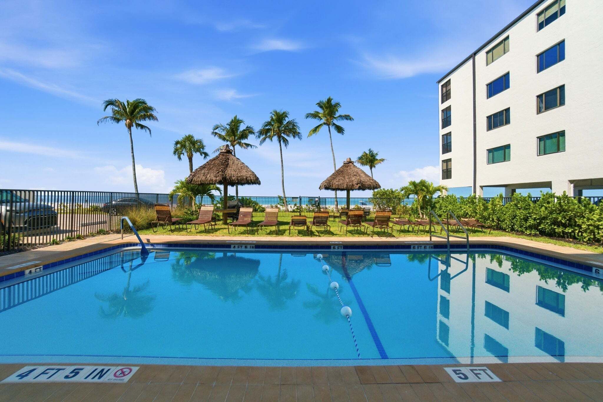 A swimming pool with a 4 FT 5 IN depth marker, surrounded by lounge chairs and palm trees, with a building in the background.