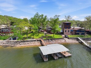 A riverside dock with a covered seating area, a stone retaining wall, and residential buildings in the background. - Historic Harbor - Timeless, Location & 2 Slips! (Lake Ozark)