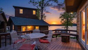 Um deck de madeira com mesa e cadeiras redondas, com vista para um corpo de água ao pôr do sol.