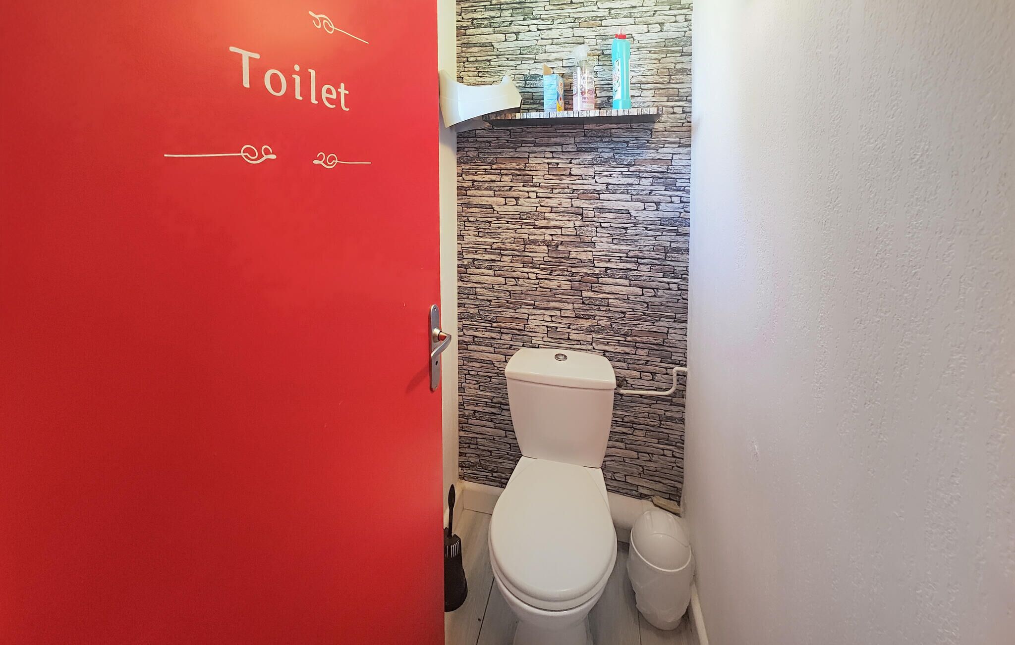 A red door with 'Toilet' written on it, a white toilet, a stone-patterned wall, and a shelf with toiletries.