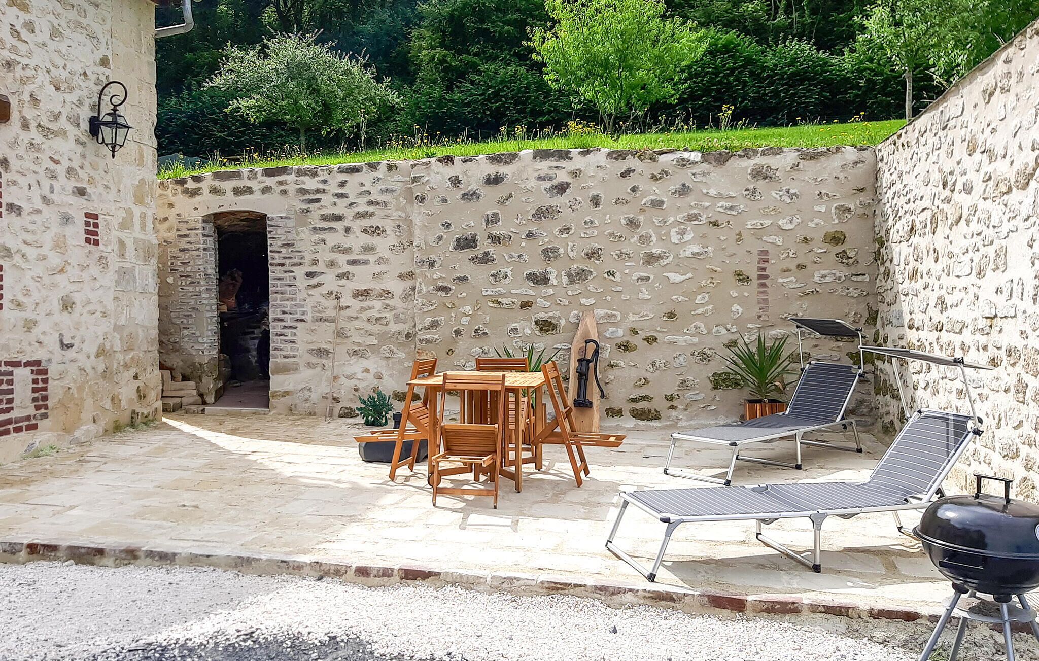 A stone-walled patio with a table and chairs, a grill, and a chair with a sunshade.