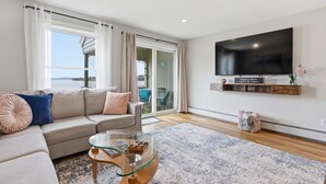 Ein Wohnzimmer mit einer Couch, einem Glastisch, einem an der Wand montierten Fernseher und Blick ins Freie.