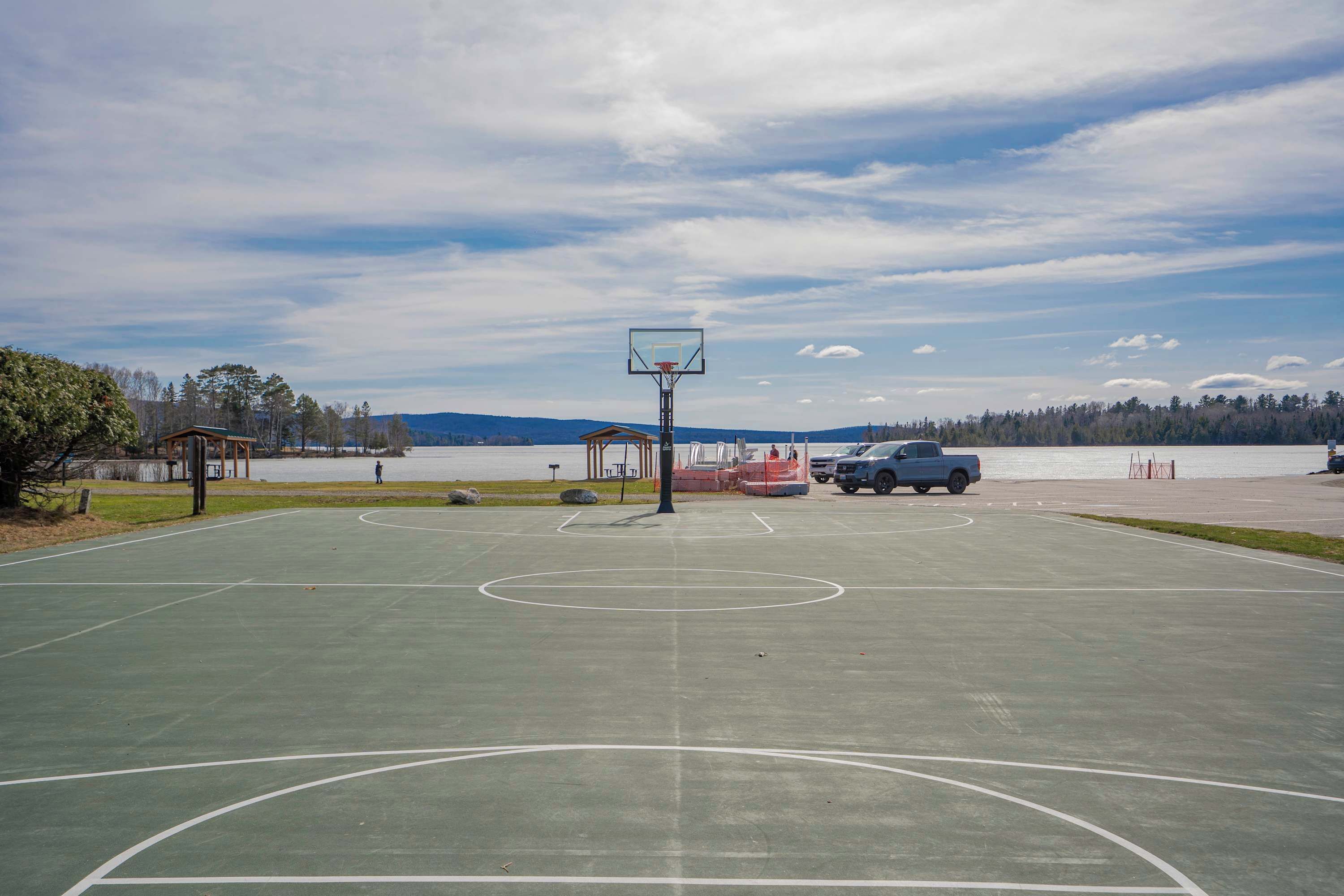 Een basketbalveld buiten met een net, een blauwe vrachtwagen en een meer op de achtergrond.