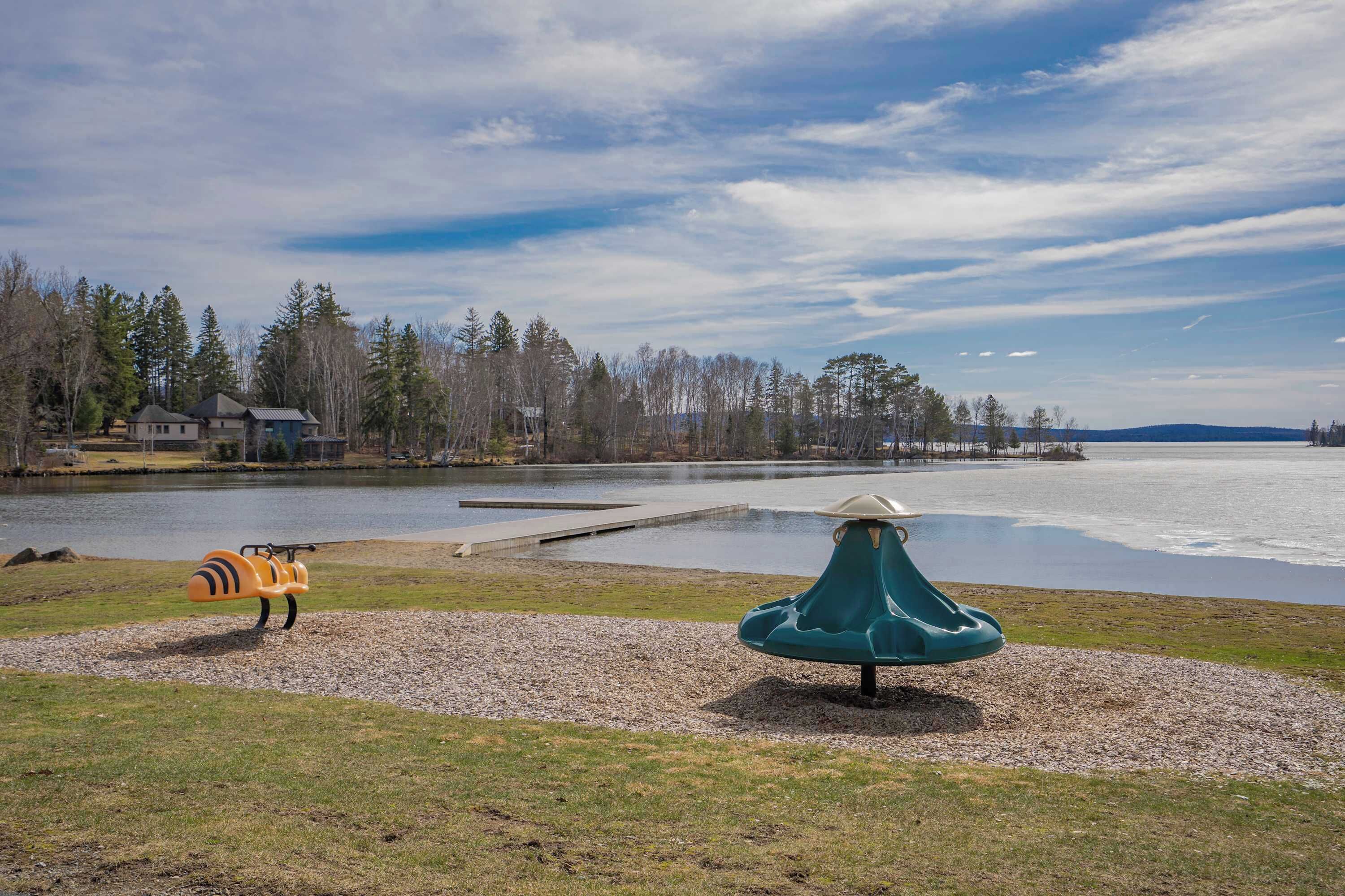 A lakeside park with playground equipment, a dock, and houses in the background.