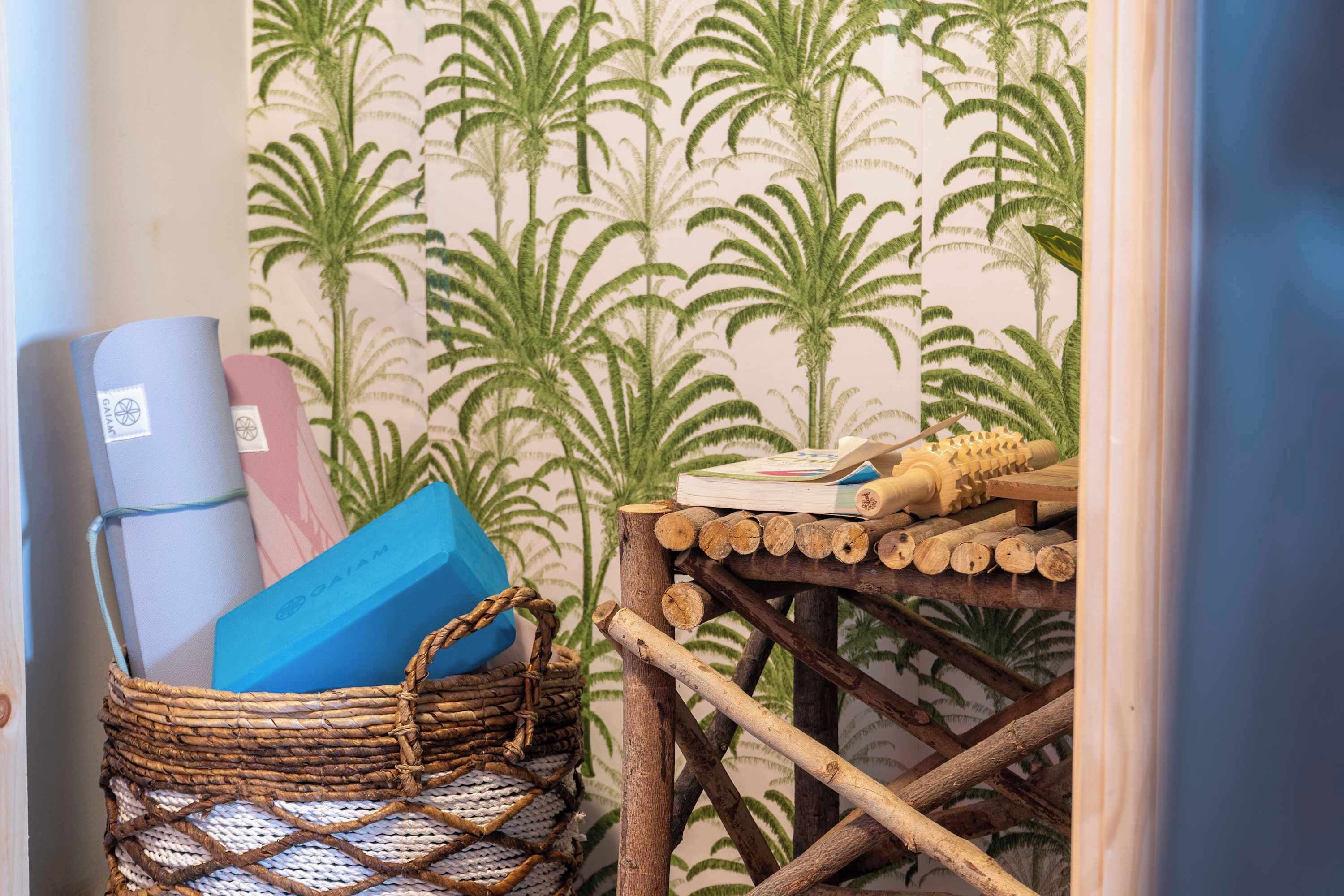 Une pièce avec un papier peint à motifs, un panier en osier rempli de livres et une petite table en bois sur laquelle se trouvent un livre et un pinceau.