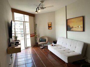 A living room with a sofa, a chair, a ceiling fan, a painting, and a window with a view of buildings. - Oceanfront apartment in Barra da Tijuca (Rio de Janeiro)
