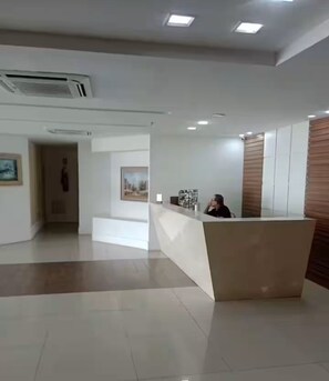 A modern reception area with a reception desk, a person seated behind it, and a hallway leading to another room. - Oceanfront apartment in Barra da Tijuca (Rio de Janeiro)