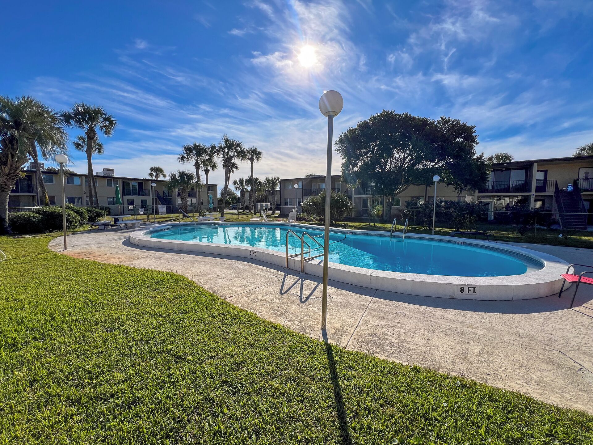 Piscina con una profundidad de 1.8 metros, rodeada de césped, palmeras y edificios de apartamentos.