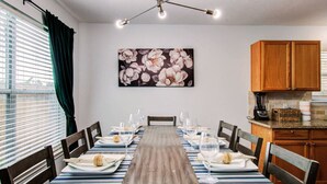 A dining room with a long table set for six, wooden chairs, a modern chandelier, and a kitchen area with wooden cabinets. - Empress Modern Haven (Houston)
