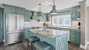 A kitchen with teal cabinetry, a stainless steel refrigerator, pendant lighting, and a marble island with stools. - Reelaxing: Oceanfront Home in Yaupon Beach, Oak Island, Perfect for 9 guests! (Oak Island)