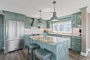 A kitchen with teal cabinetry, a stainless steel refrigerator, pendant lighting, and a marble island with stools.
