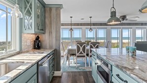A kitchen with a large island, a dining area with a view, and a ceiling fan. - Reelaxing: Oceanfront Home in Yaupon Beach, Oak Island, Perfect for 9 guests! (Oak Island)