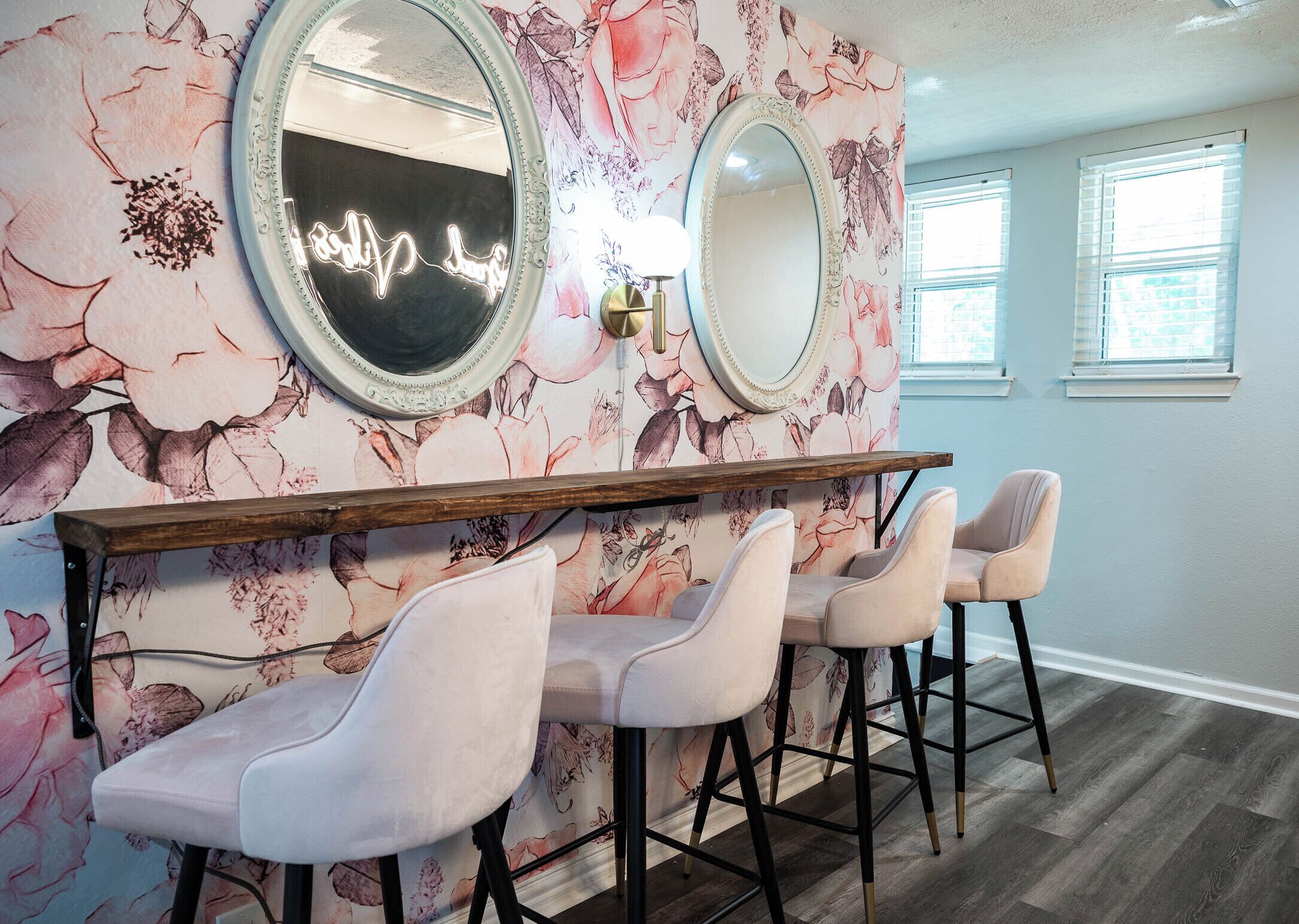 A room with floral wallpaper, two large mirrors, a wooden bar, and three chairs.