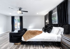 Un dormitorio moderno con una cama, sillones tapizados en negro, un ventilador de techo y una ventana con cortinas.