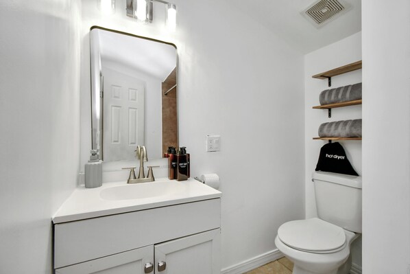 A bathroom with a white sink, mirror, and toilet. There are shelves above the toilet with towels and a black cap labeled 'hardyrier'. - Empress Sunlit Villa (Cypress)