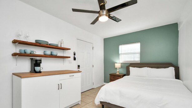 A bedroom with a bed, a ceiling fan, a coffee maker, and a window.