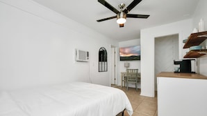 A bedroom with a bed, a ceiling fan, a desk with a coffee machine, a chair, and a painting on the wall.