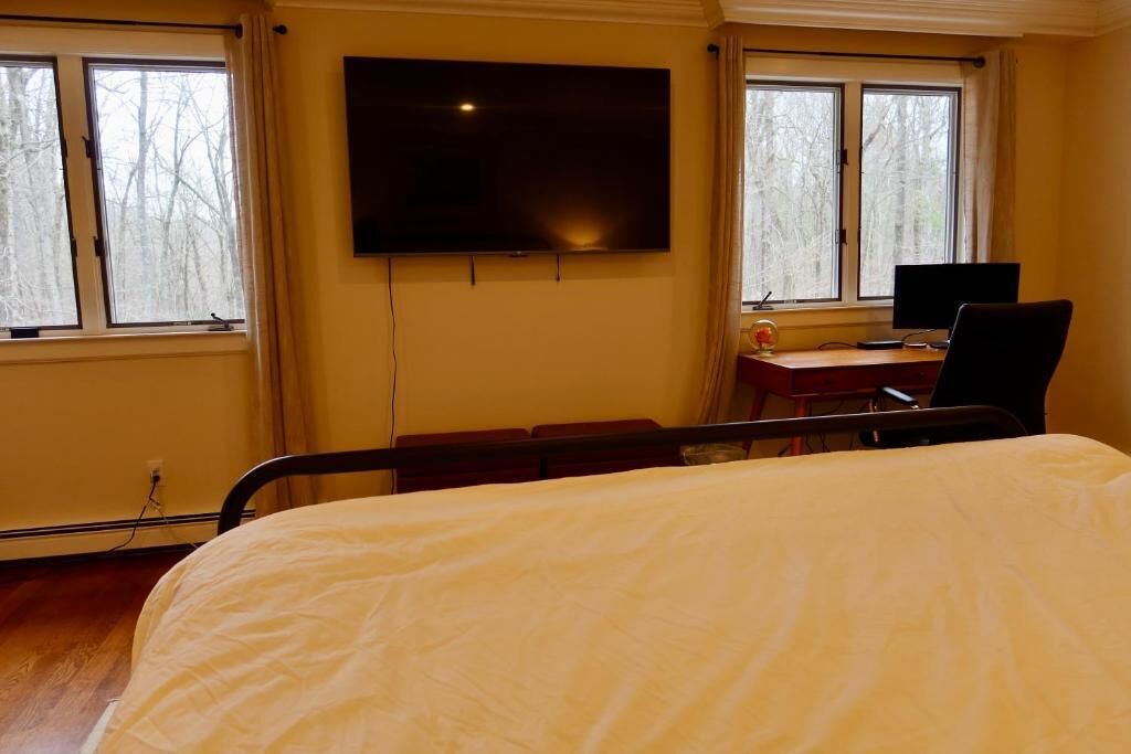 Una habitación de hospital con una cama, un escritorio con computadora, un televisor y ventanas que dan a árboles.
