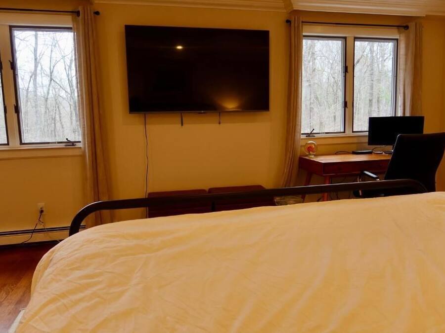 Una habitación de hospital con una cama, un escritorio con computadora, un televisor y ventanas que dan a árboles.