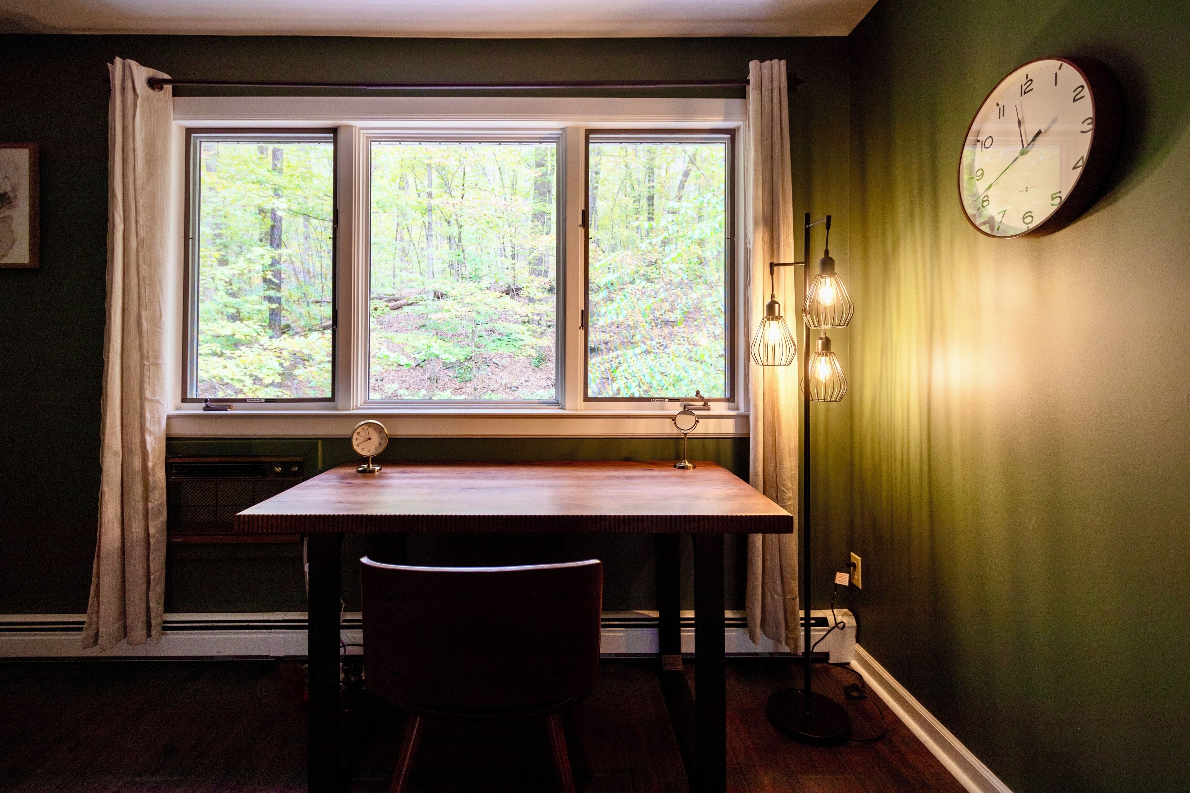 A room with a window, a clock, and a table with a chair.