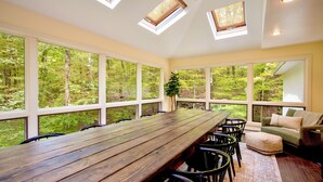 A room with a long wooden table, multiple chairs, a sofa, and large windows offering a forest view.