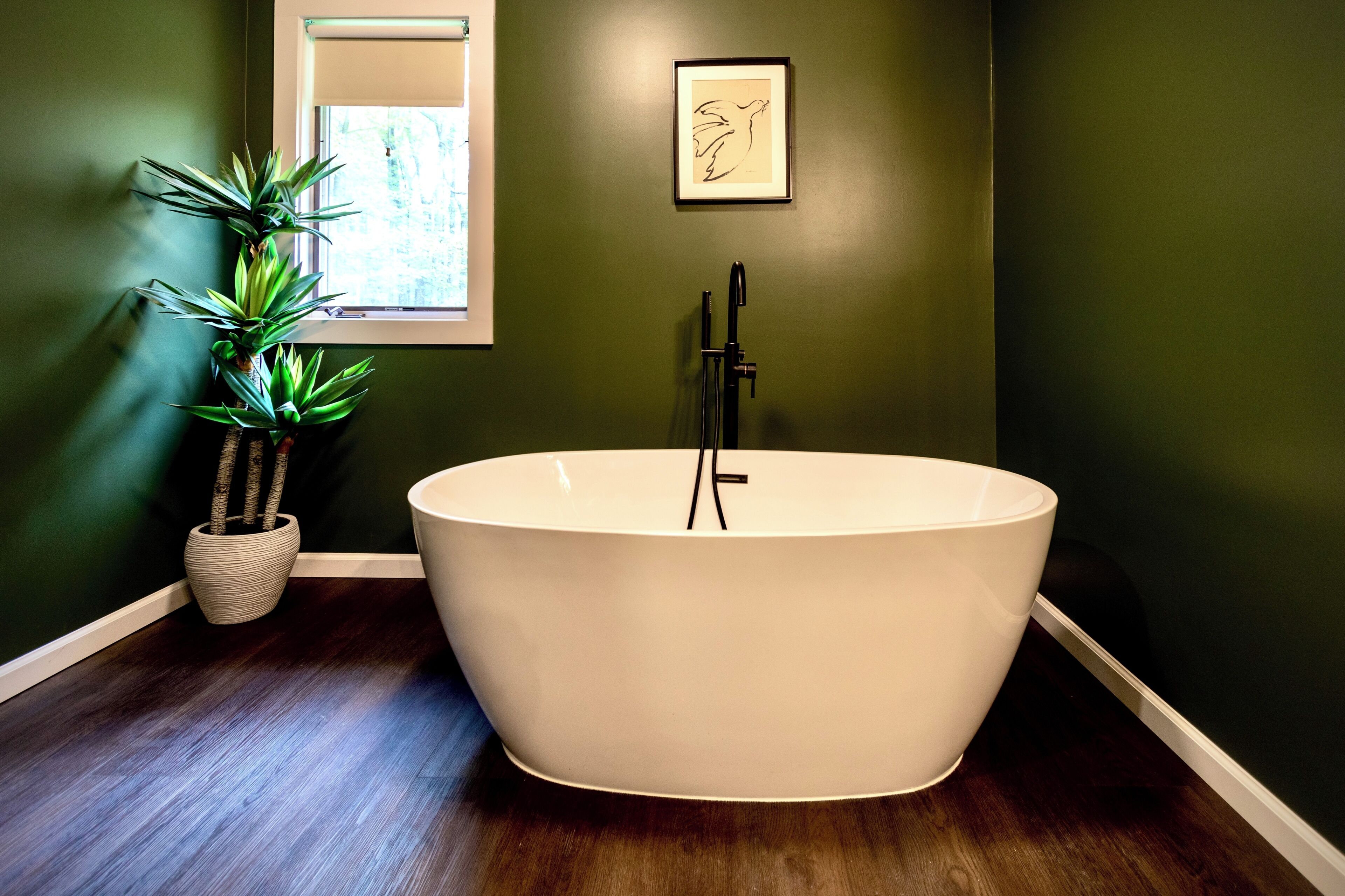 A freestanding bathtub in a room with dark green walls, a window with blinds, a framed artwork, and a potted plant.