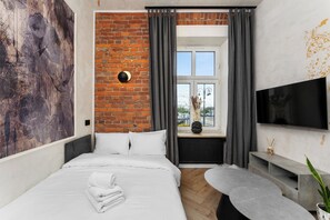 Ein Hotelzimmer mit Bett, Fernseher, Fenster mit Vorhängen, einer Ziegelwand und einer Pflanze.