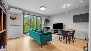 Ein modernes Wohnzimmer mit einem türkisblauen Sofa, Holzböden, einem Essbereich mit Stühlen und Tisch, einem Fernseher und einem großen Fenster mit Blick auf Grünflächen.