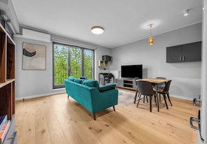 Ein modernes Wohnzimmer mit einem türkisblauen Sofa, Holzböden, einem Essbereich mit Stühlen und Tisch, einem Fernseher und einem großen Fenster mit Blick auf Grünflächen.