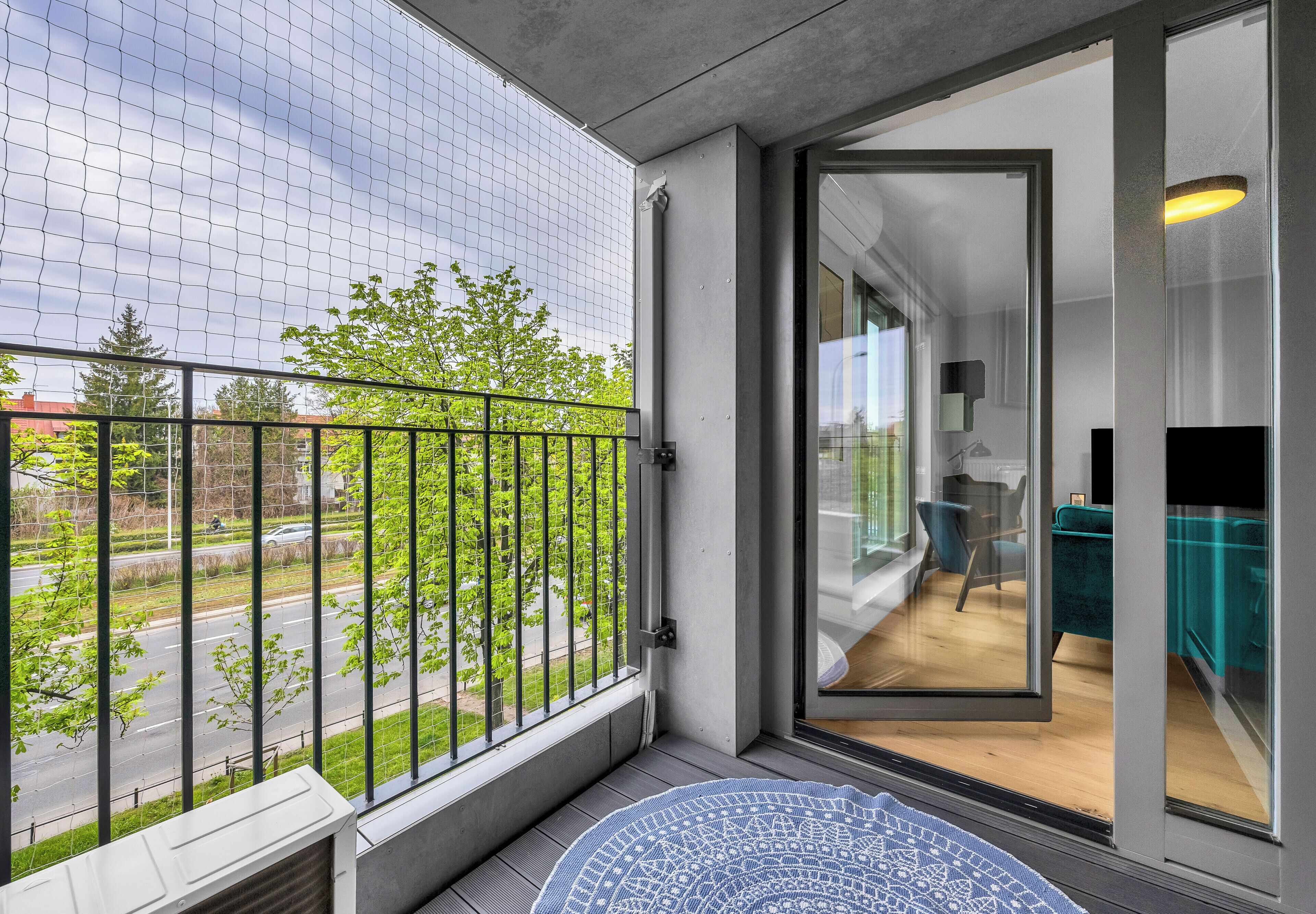 Un balcon donnant sur une route et des arbres, une porte-fenêtre coulissante qui mène à une pièce avec un canapé turquoise et un plancher en bois.