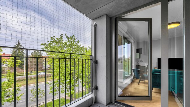 Ein Balkon mit Blick auf eine Straße und Bäume, eine Schiebetür zu einem Raum mit einem türkisblauen Sofa und Holzboden.