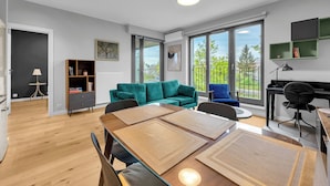 Ein modernes Wohnzimmer mit Essbereich, einem grünen Sofa, einem Schreibtisch und großen Fenstern mit Blick auf Bäume.