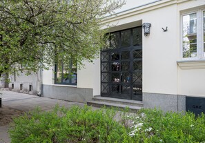 Un bâtiment doté d’une grande fenêtre, d’une porte ornée d’un motif en croix et d’un petit jardin avec des arbustes et un arbre.