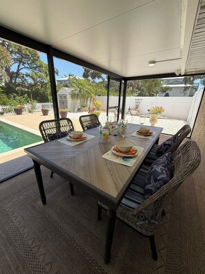Eine überdachte Terrasse mit Esstisch, gedeckt für vier Personen, umgeben von Korbstühlen, einem Pool und einem weißen Zaun.